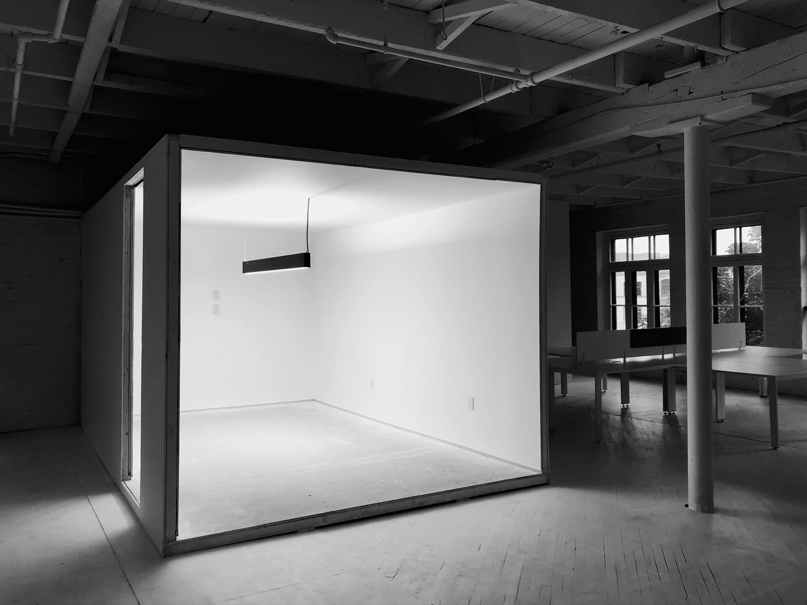 Une pièce blanche entièrement close construite au cœur d’un loft industriel sombre, éclairée par un seul luminaire linéaire suspendu.